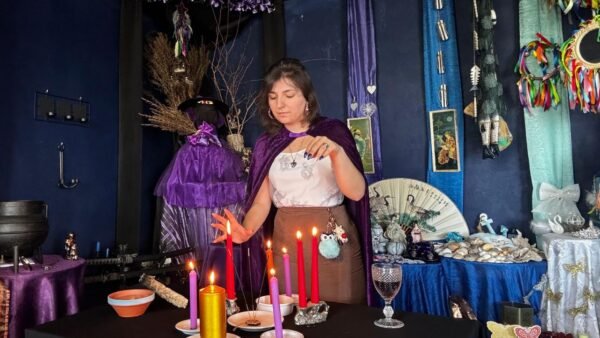 Mica Mihaylova em frente a um altar mágico com velas acesas de várias cores, incensos e objetos espirituais, vestida com capa roxa em ambiente azul ritualístico