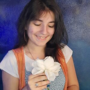 Mica Mihaylova a segurar uma flor branca com as duas mãos e a sorrir suavemente, com fundo azul texturizado