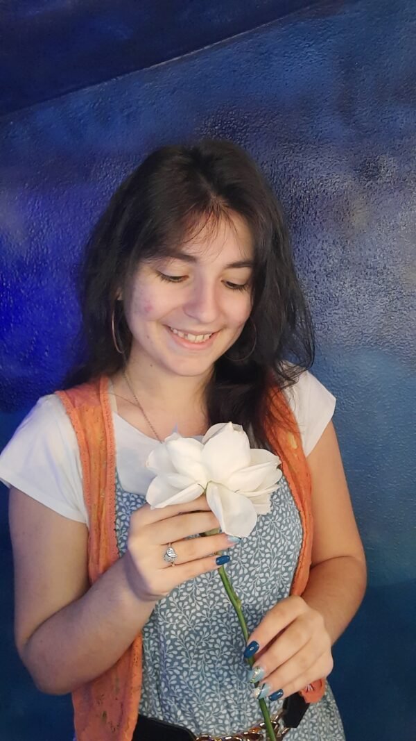 Mica Mihaylova a segurar uma flor branca com as duas mãos e a sorrir suavemente, com fundo azul texturizado