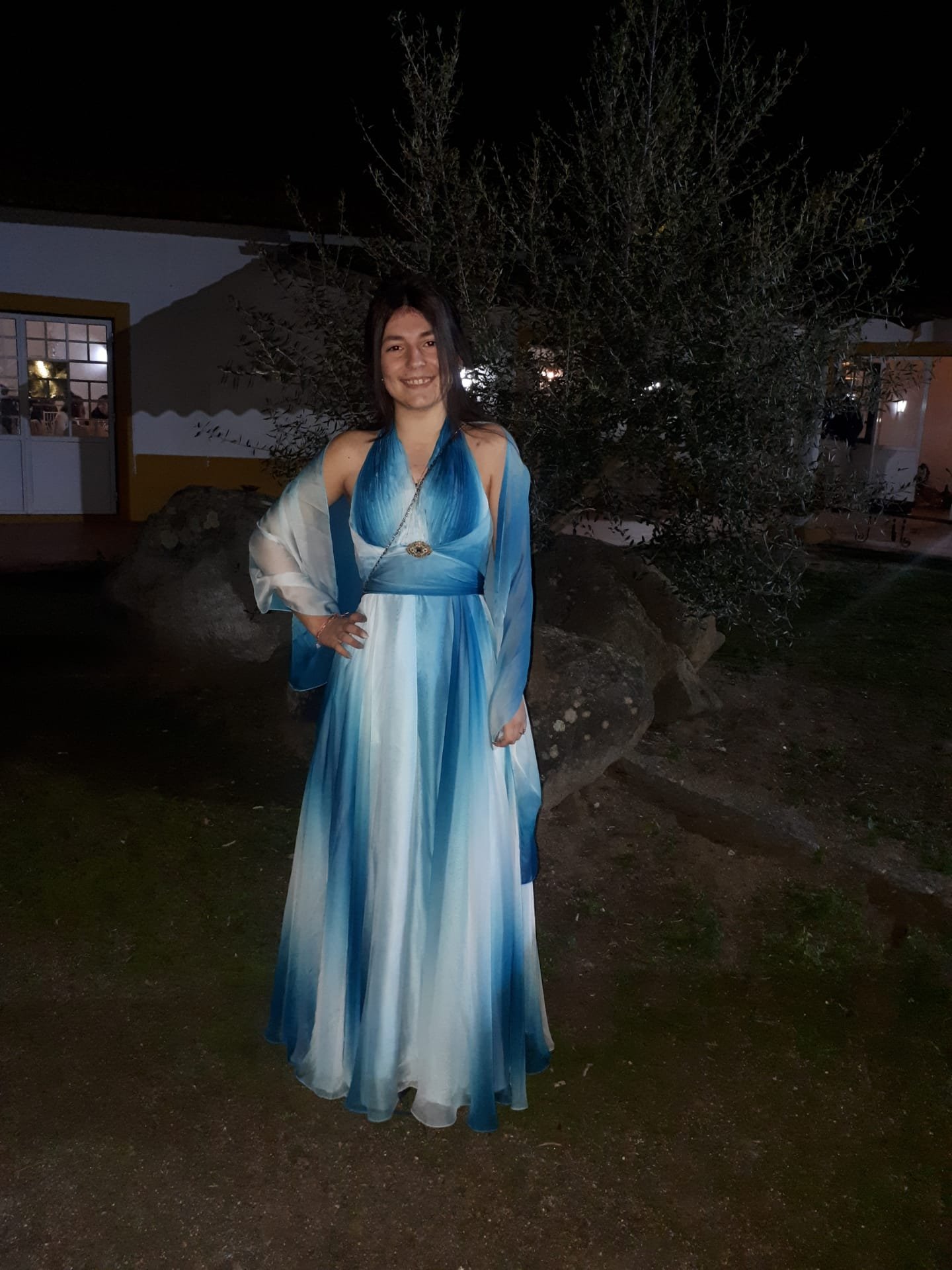 Mica Mihaylova em pé com um vestido azul e branco esvoaçante, sorridente e em ambiente noturno com vegetação ao fundo