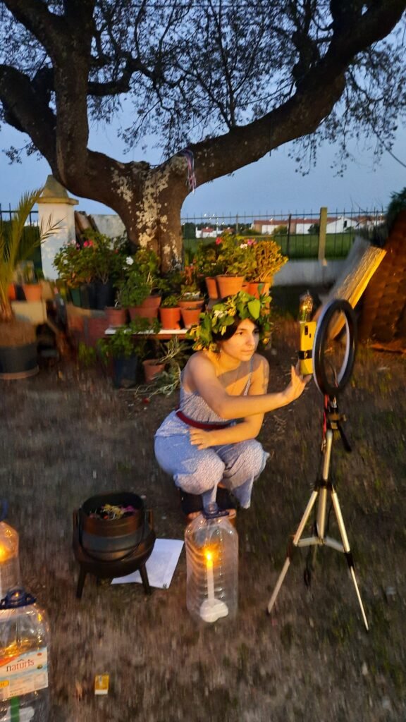 Mica Mihaylova de cócoras com coroa de folhas e flores, em frente a um tripé com luz e caldeirão ritual, rodeada de plantas e velas